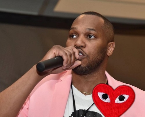 Photo of Kenya Hutton standing holding a microphone in his right hand. Wearing a pink blazer.
