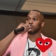 Photo of Kenya Hutton standing holding a microphone in his right hand. Wearing a pink blazer.