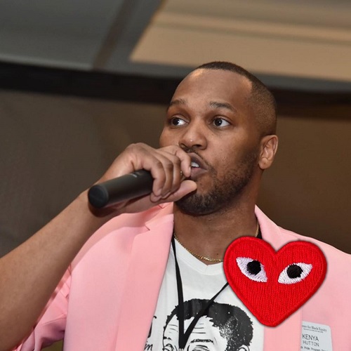 Photo of Kenya Hutton standing holding a microphone in his right hand. Wearing a pink blazer.